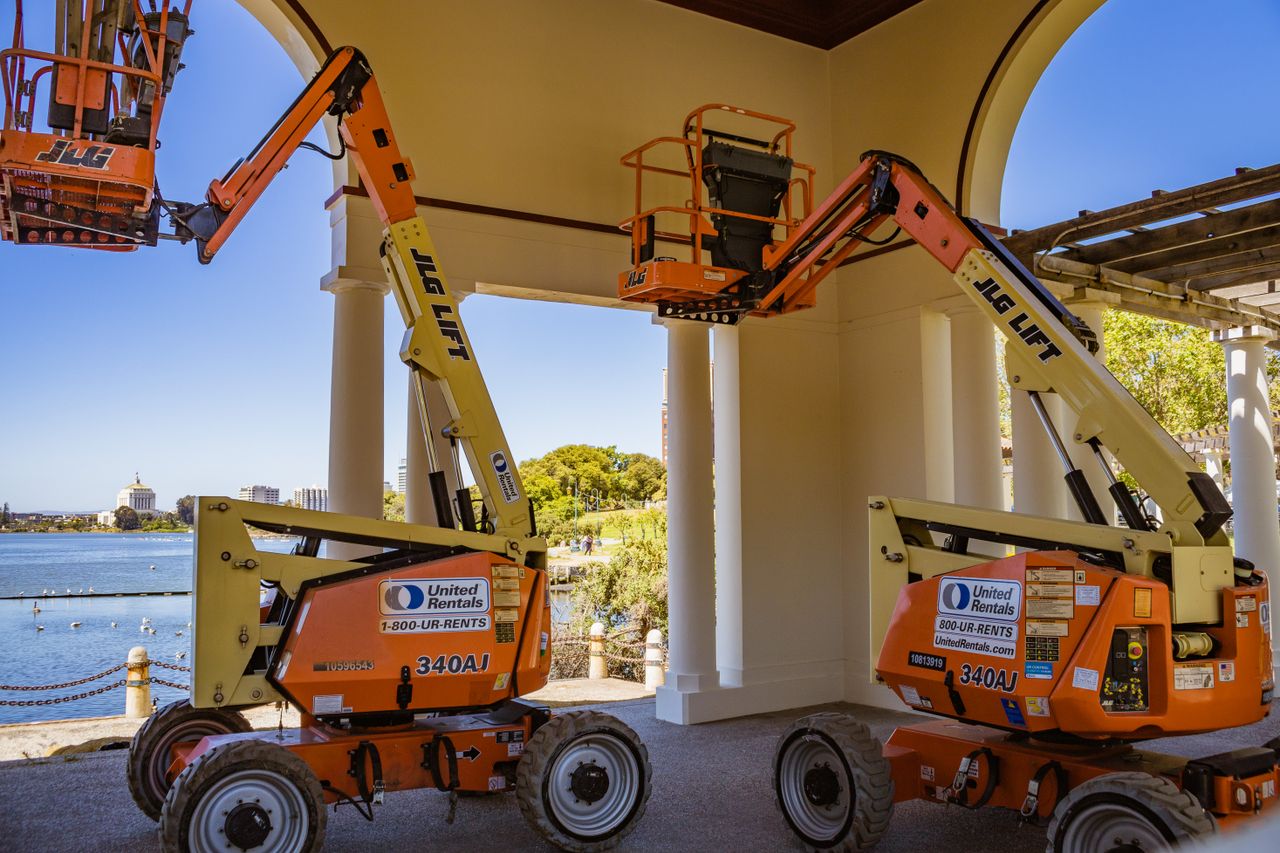 story-01 Orange construction lifts positioned near a lakeside with clear sky and architecture.